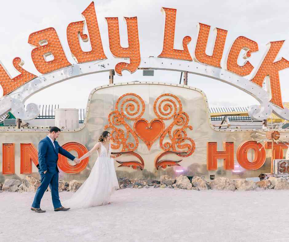 Couple de mariés marchant main dans la main devant l’enseigne vintage Lady Luck au Neon Museum de Las Vegas, photo Pretty Day.