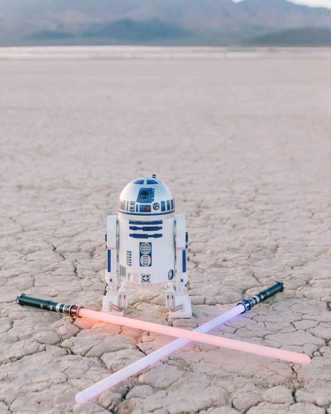 Figurine de R2-D2 posée dans le désert, entourée de deux sabres laser croisés au sol.