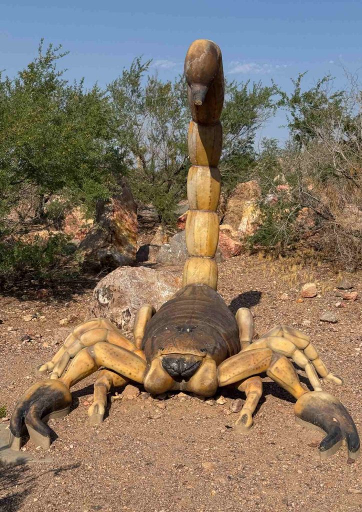 Sculpture de scorpion avec la queue dressée sur un sol de gravier, dans un paysage désertique.