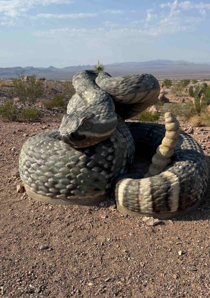 Sculpture d’un grand serpent enroulé sur un sol de gravier, dans un paysage désertique.