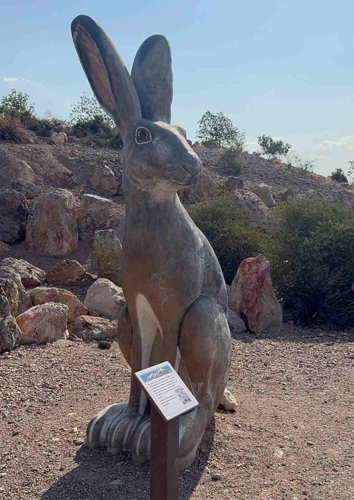 Grande sculpture de lapin assis près d’un panneau d’information, sur un terrain caillouteux.