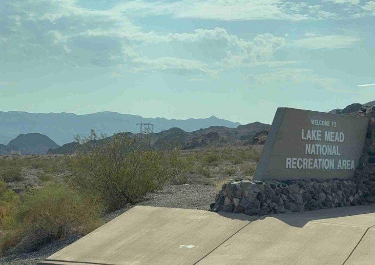 Panneau d’entrée indiquant “Lake Mead National Recreation Area” au bord d’une route, avec collines au loin.
