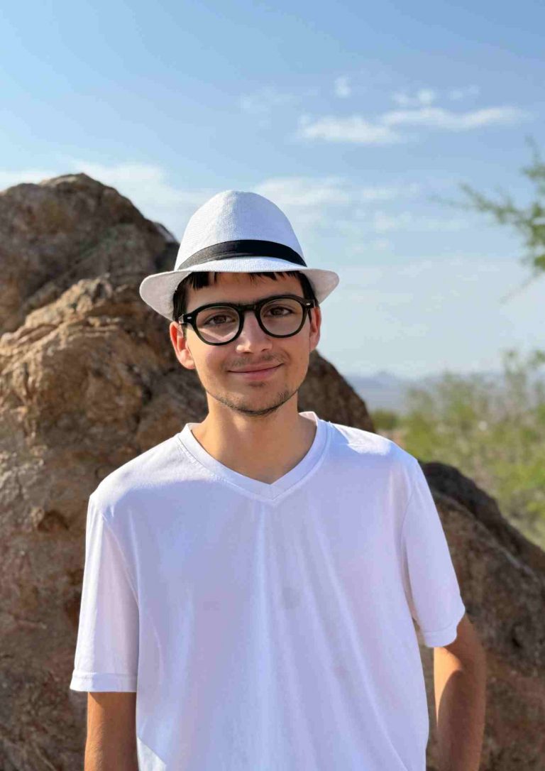 Personne portant un chapeau clair et des lunettes, debout devant un gros rocher.